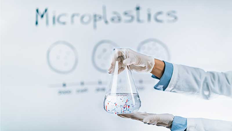 Blog Ambiental • Cientista segura frasco com microplásticos em líquido, com fundo explicativo sobre o tema