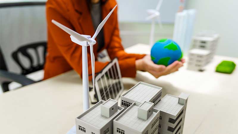Blog Ambiental • Mulher segura planeta em miniatura ao lado de maquetes de prédios, turbinas eólicas e placas solares