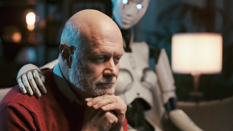 Homem maduro, calvo e com expressão pensativa, é confortado por um robô humanoide branco, que apoia a mão em seu ombro em um ambiente doméstico iluminado.