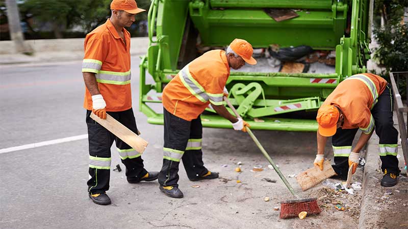Três agentes ambientais uniformizados limpando resíduos sólidos em frente a caminhão de coleta de lixo