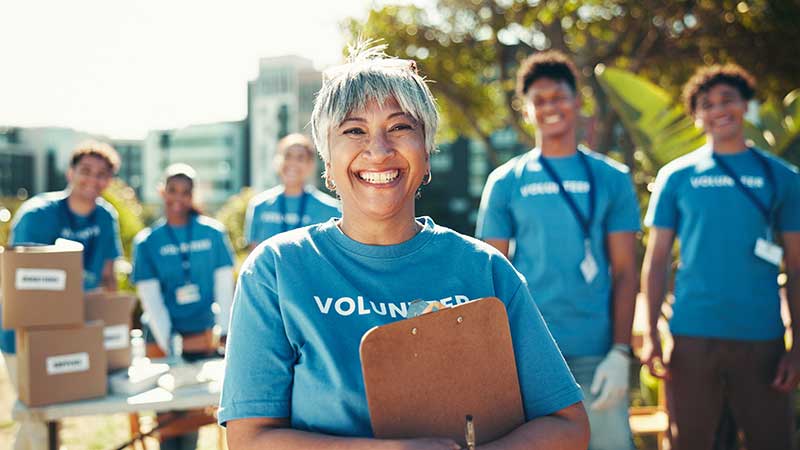 Grupo de voluntários sorridentes com camisetas azuis, liderados por mulher segurando uma prancheta em ação comunitária ao ar livre