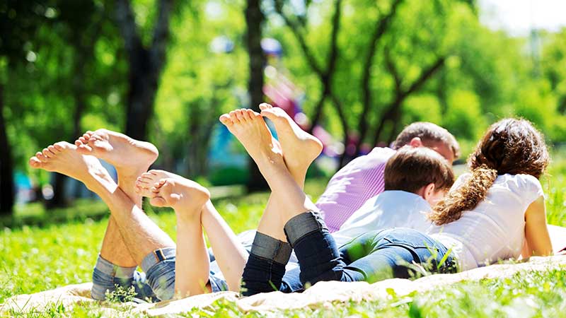 Família deitada na grama em dia ensolarado em parque natural, desfrutando de momentos de relaxamento e bem-estar na natureza.