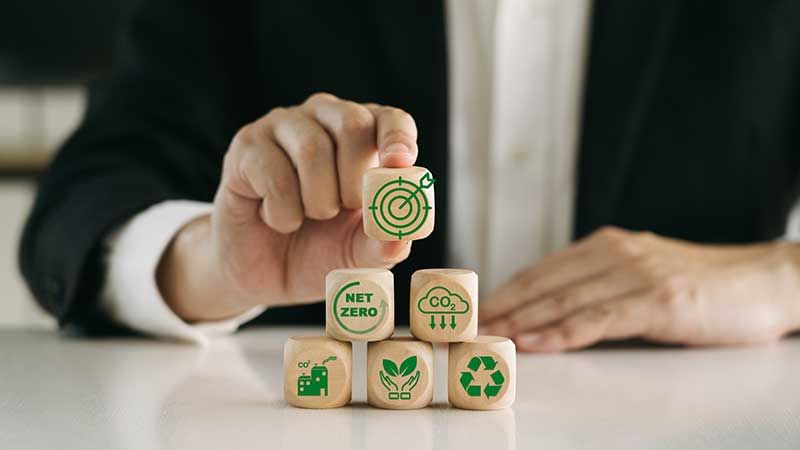 Mão organizando blocos de madeira com ícones de sustentabilidade, representando economia verde, reciclagem e redução de CO₂.