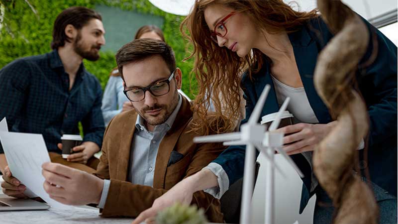 Profissionais analisam documentos e modelos de turbinas eólicas em uma reunião sobre sustentabilidade e energia renovável.