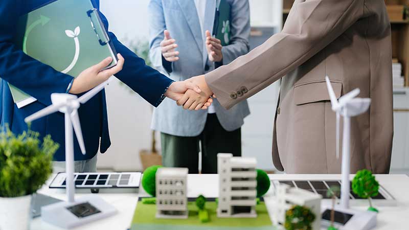 Aperto de mãos entre profissionais discutindo soluções ambientais, com modelos sustentáveis e turbinas eólicas na mesa.