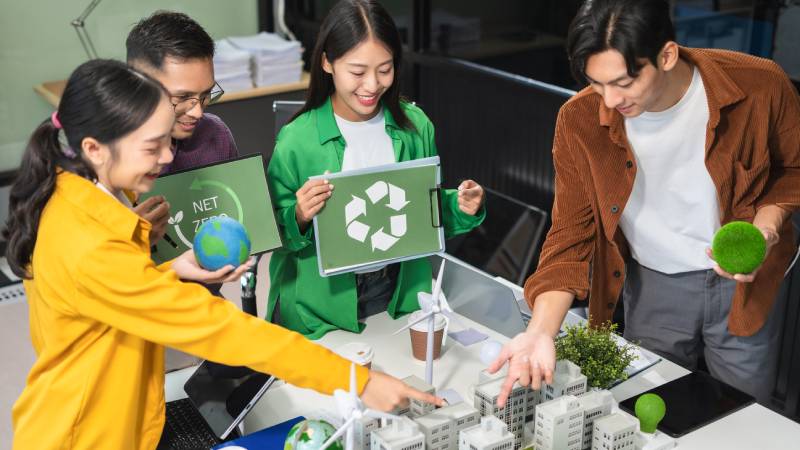 Blog Ambiental • Jovens profissionais discutem projetos sustentáveis sobre maquete de cidade, segurando ícones de reciclagem e planeta Terra, representando ou uso do PDI na economia circular.
