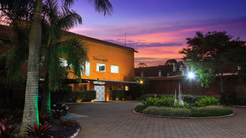 Fachada do Hotel Terras Altas iluminada ao entardecer, com céu colorido e paisagismo na entrada.